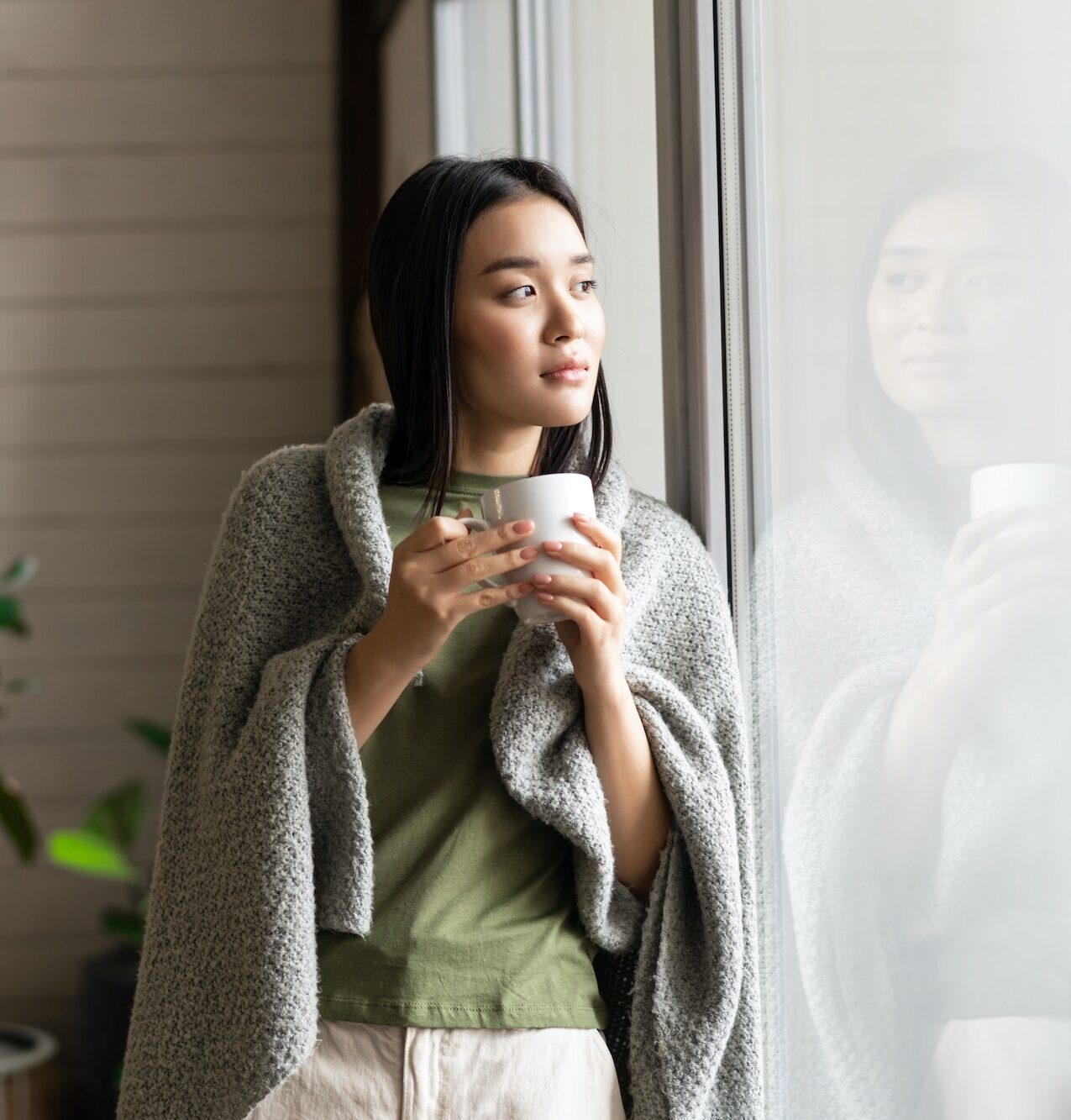 Beautiful asian woman wrapped in blanket, leaning on window and looking outside, drinking hot tea
